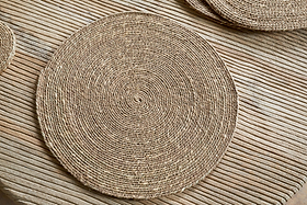 The nkuku Keso Woven Placemats - Brown (Set of 4) rest on a slatted wooden surface, with another placemat and handwoven coasters in view, adding minimalist style to your tableware.