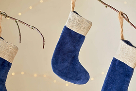 Three Karru Cotton Velvet Mini Stockings in indigo by nkuku (set of 3) hang from bare branches with burlap cuffs and soft string lights, creating a cozy, festive scene against a light background.