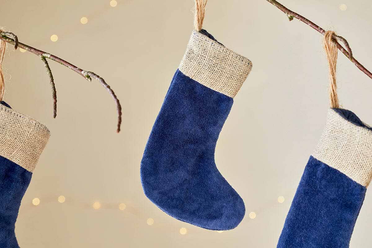 Three Karru Cotton Velvet Mini Stockings in indigo by nkuku (set of 3) hang from bare branches with burlap cuffs and soft string lights, creating a cozy, festive scene against a light background.