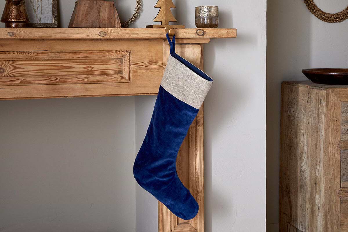 The nkuku Karru Cotton Velvet Stocking in Indigo, featuring a beige cuff, hangs from a rustic wooden mantel surrounded by festive décor like a small tree and candle holders, adding charm to the cozy room.
