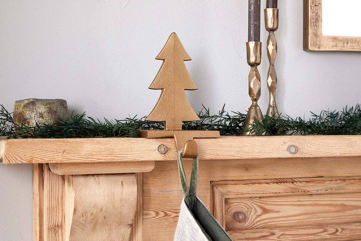 A wooden mantelpiece features green garland, a gold metal tree figurine, two gold candlesticks with brown candles, and a rustic gold candle holder. A stocking hangs below on nkuku's Kotta Stocking Tree Holder in antique brass.