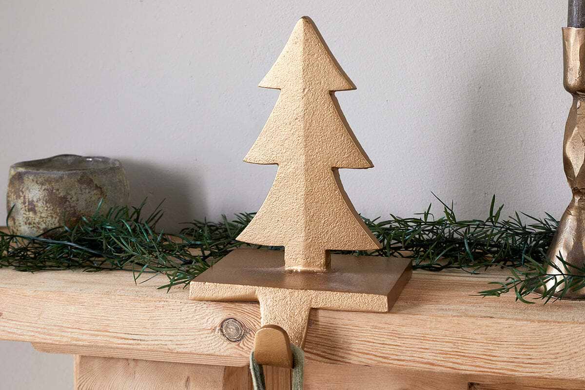The Kotta Stocking Tree Holder in antique brass by nkuku sits on a wooden mantel with green garland. The brass hook hangs over the edge for stockings, and a small candleholder and candlestick are partially visible nearby.