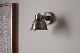 The nkuku Kasai Brass Spotlight in Antique Brass is mounted on a light-colored, textured wall, with part of a framed fabric artwork visible on the left.