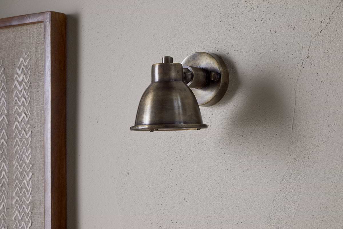 The nkuku Kasai Brass Spotlight in Antique Brass is mounted on a light-colored, textured wall, with part of a framed fabric artwork visible on the left.