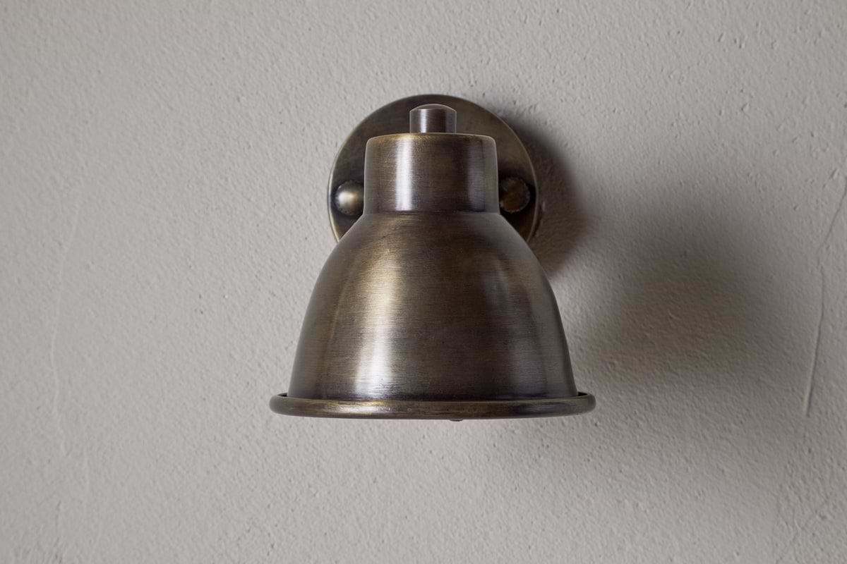 The Kasai Brass Spotlight by nkuku features an antique brass finish and dome-shaped shade, mounted on a light gray textured wall. This adjustable fixture brings vintage industrial style and character to any space.