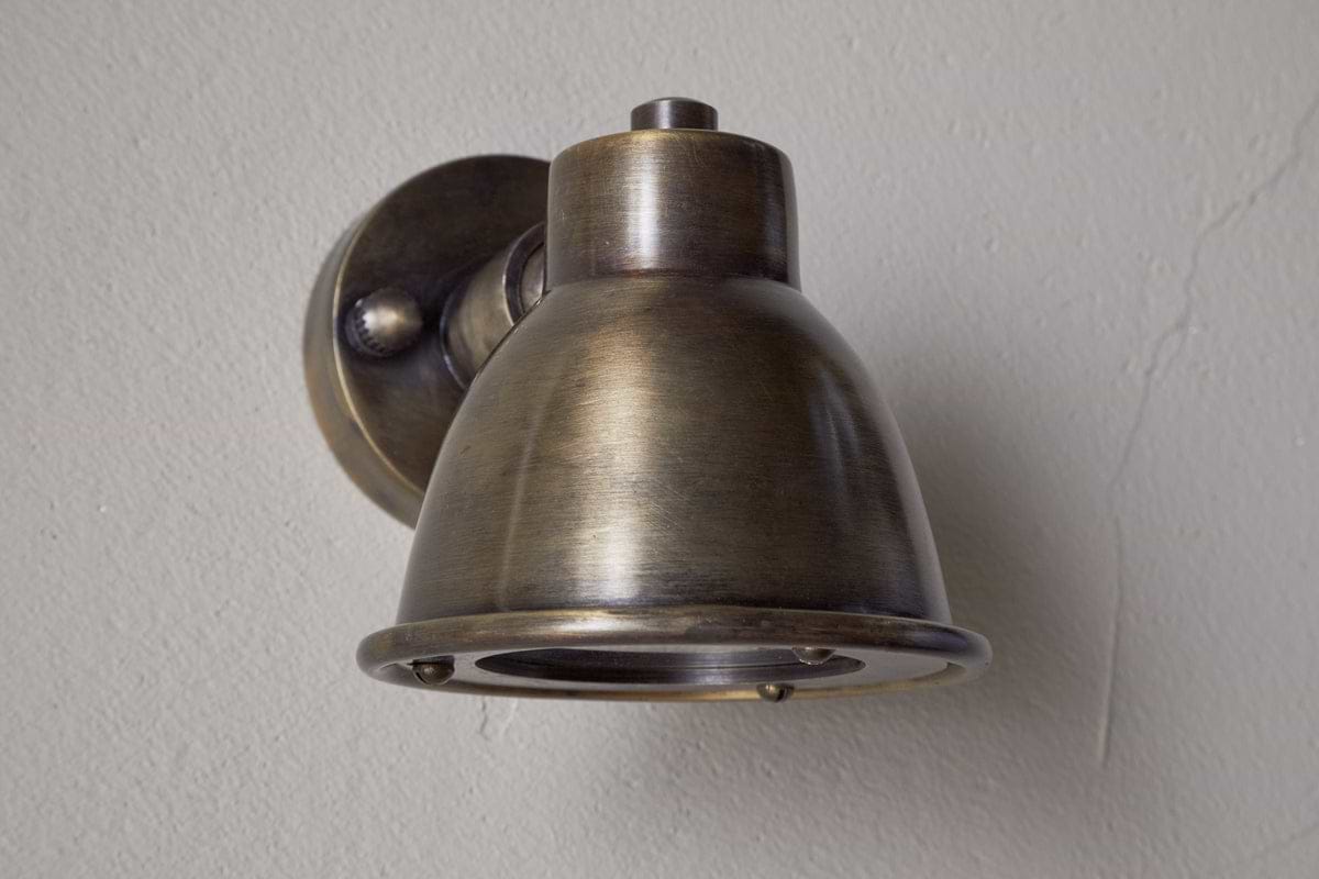 The Kasai Brass Spotlight by nkuku features a dome-shaped, industrial-style antique brass shade on a round wall plate, mounted to a plain light-colored wall for a rustic vintage look.