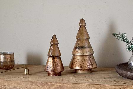 Two nkuku Konara Glass Trees in gold crackle add a festive touch on a wooden surface. The larger tree sits on the right beside a gold bell, glass candle holder, and vase with greenery against a plain wall.