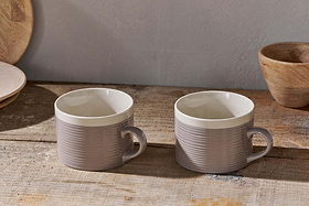 The nkuku Kai Mugs - Grey (Set of 2) feature a contemporary ribbed design and white interiors, displayed on a rustic wooden surface with wooden bowls and plates in the background for a cozy, natural kitchen vibe.