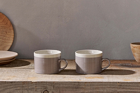 The Kai Mugs - Grey (Set of 2) by nkuku, featuring contemporary ribbed detail, rest on a rustic wooden surface with light brown wooden plates and a bowl behind them, all softly lit for a cozy ambiance.