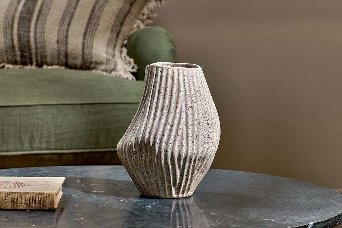 The Kalai Ceramic Organic Shape Vase - Small by nkuku, in textured beige with vertical ridges and hand-shaped in Portugal, rests on a dark marble table. A green upholstered chair with a striped pillow adds to the cozy, modern atmosphere.