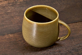 The nkuku Kalini Mug - Green (Extra Large) has a textured, speckled olive finish, a lighter tan rim, and a curved handle. It sits on a rustic dark brown wooden surface—perfect for enjoying your favorite hot drinks.