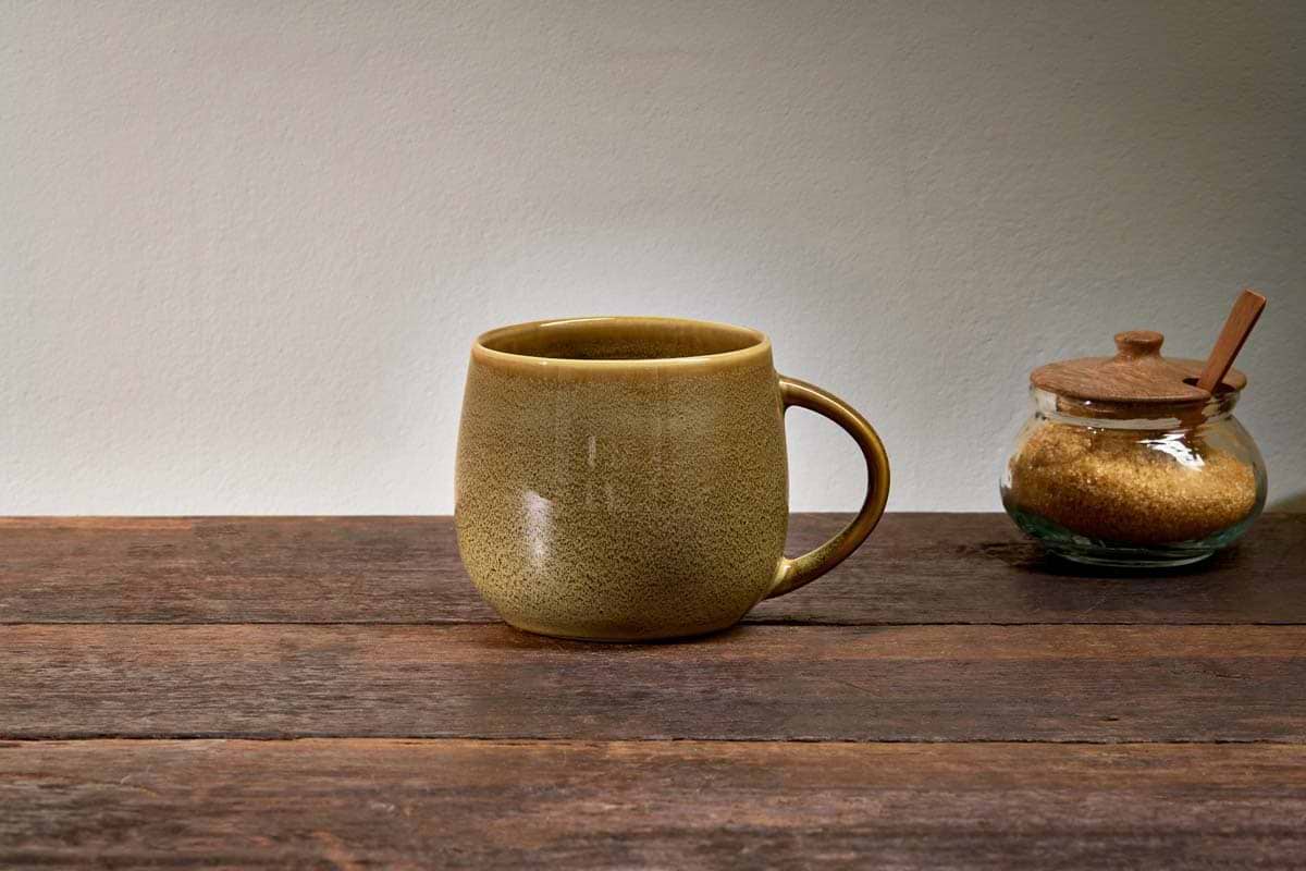 An extra large, green Kalini Mug by nkuku sits on a wooden table beside a glass jar of brown sugar and a wooden spoon.