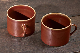 A set of two large, kiln-fired Kalini Mugs - Amber by nkuku with deep red bodies and light brown rims rests on a dark, weathered wooden surface; the left mug sits closer and faces left, the right mug faces right.