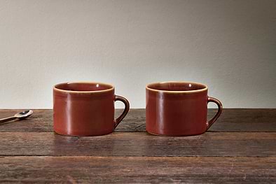 Two kiln-fired nkuku Kalini Mugs - Amber - Large (Set of 2) with handles rest side by side on a rustic wooden table. A small metal spoon with a dark handle lies to the left, set against a light beige background for a simple, cozy vibe.