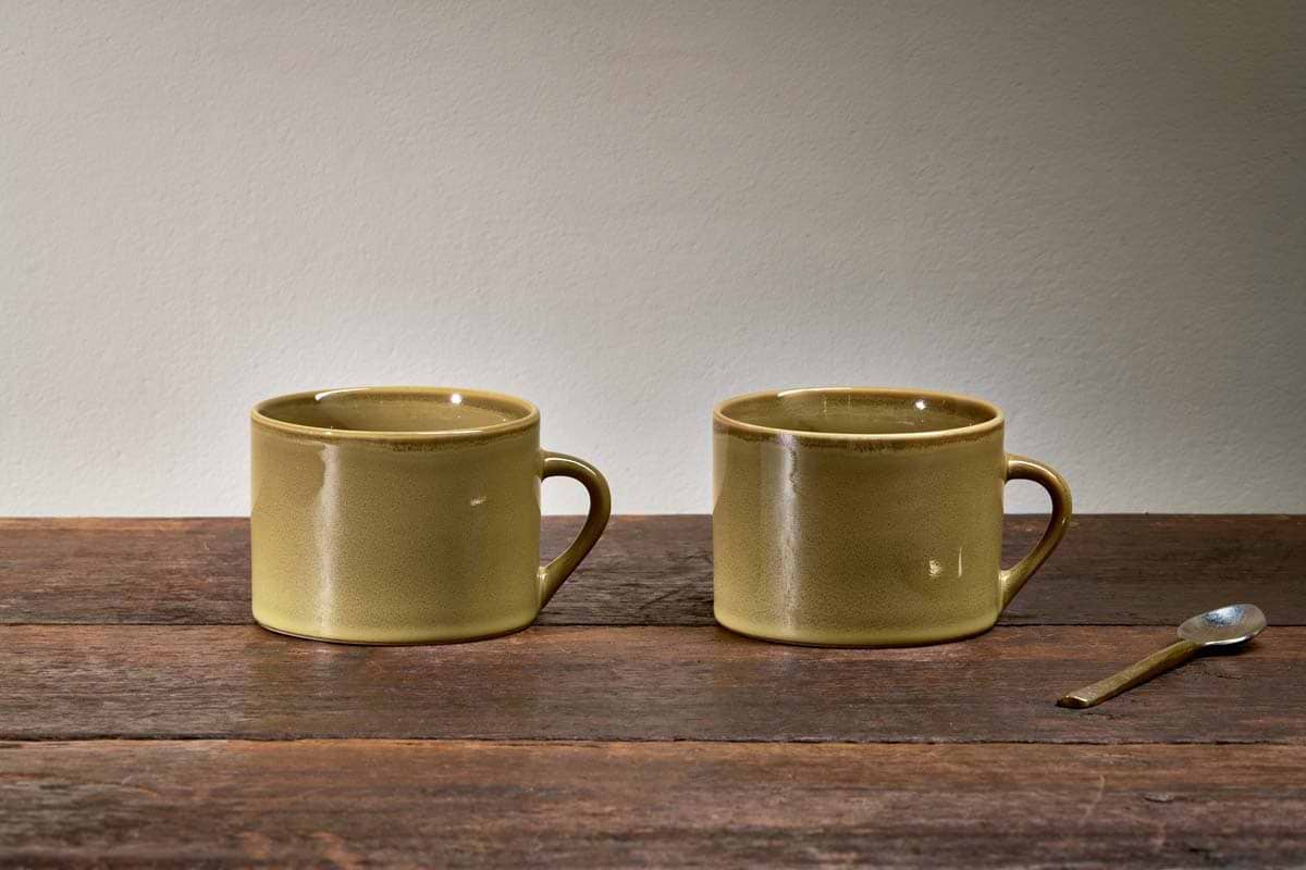 Two large green Kalini Mugs by nkuku are placed on a wooden table, accompanied by a metal spoon.