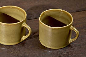 Two Kalini Mugs - Green - Small (Set of 2) by nkuku sit on a dark wooden surface, featuring simple handles and a smooth, slightly speckled green finish. One mug is fully visible, while the other is partially cut off at the edge.