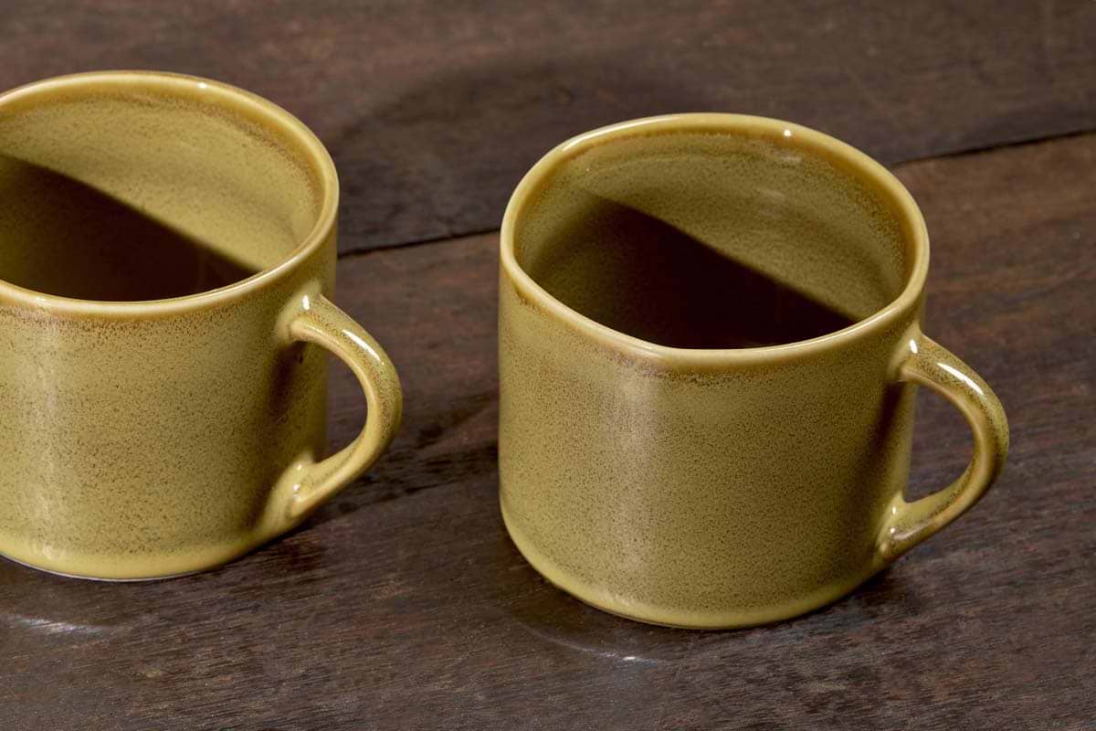 Two Kalini Mugs - Green - Small (Set of 2) by nkuku sit on a dark wooden surface, featuring simple handles and a smooth, slightly speckled green finish. One mug is fully visible, while the other is partially cut off at the edge.