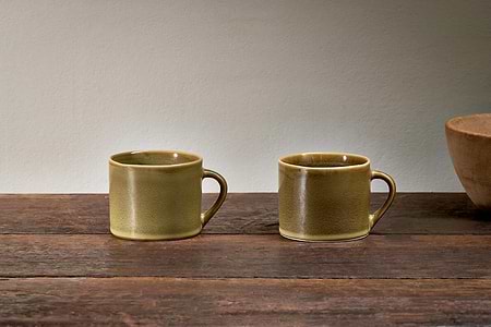 A set of two small green Kalini mugs by nkuku with handles are placed side by side on a rustic wood surface, with a brown ceramic bowl partly visible on the right and a plain, light-colored wall in the background.