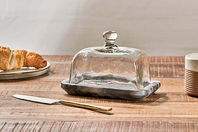 The nkuku Kallso Marble Butter Dish rests on a wooden table, its recycled glass lid with a marble base. Beside it are a gold-handled butter knife, a plate holding a croissant, and a ridged brown-and-white ceramic cup.