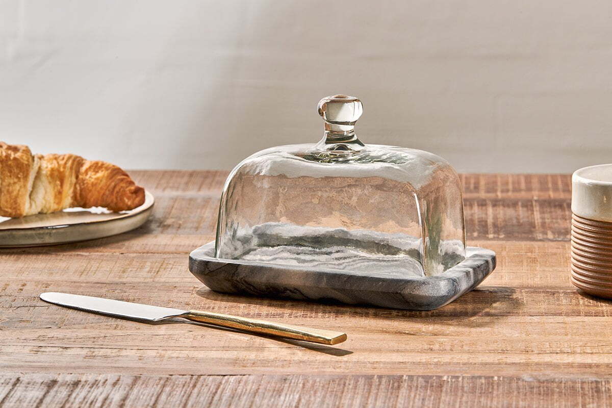 The nkuku Kallso Marble Butter Dish rests on a wooden table, its recycled glass lid with a marble base. Beside it are a gold-handled butter knife, a plate holding a croissant, and a ridged brown-and-white ceramic cup.