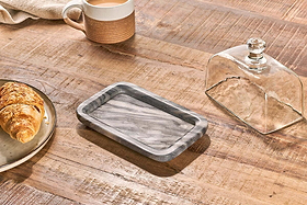 A wooden table features the nkuku Kallso Marble Butter Dish with lid, a plate holding a croissant, and a brown and white mug. Sunlight streams in, casting shadows that highlight the textures of both the items and the tabletop.