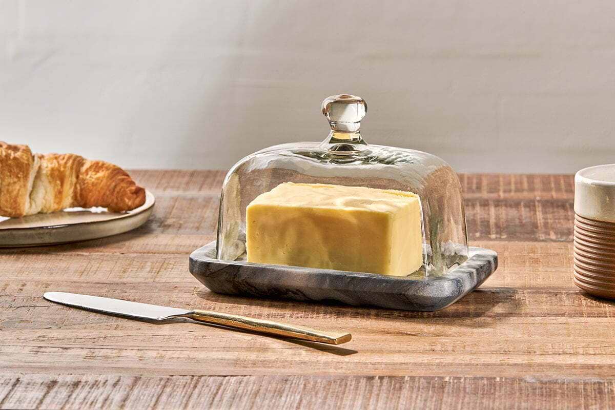 The Kallso Marble Butter Dish by nkuku holds a block of butter beneath a clear glass dome. Beside it, a gold-handled knife, striped cup, and croissant rest on a wooden table set against a neutral backdrop.