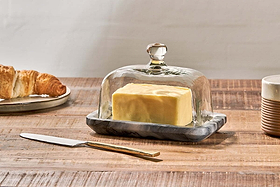 The Kallso Marble Butter Dish by nkuku holds a block of butter beneath a clear glass dome. Beside it, a gold-handled knife, striped cup, and croissant rest on a wooden table set against a neutral backdrop.