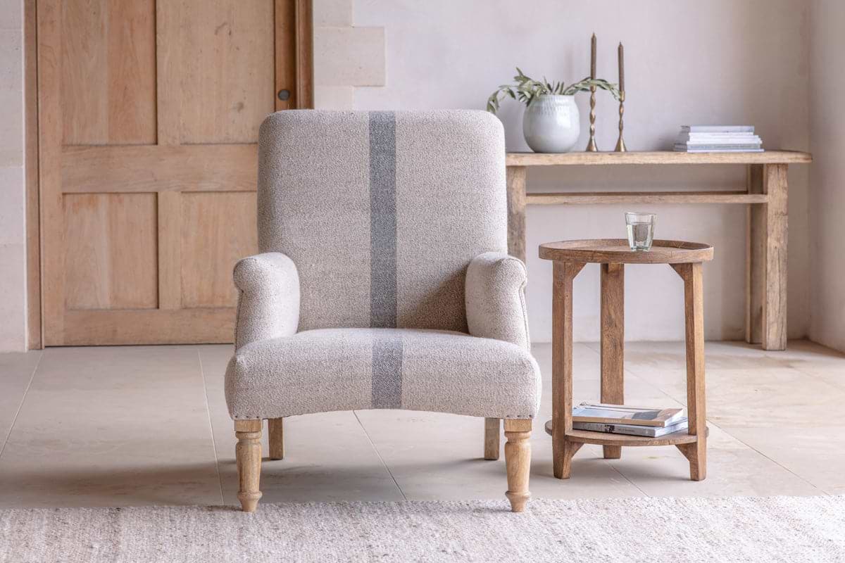 The Kallu Wool & Cotton Occasional Chair by nkuku, in natural beige with textured upholstery and gray stripes, sits beside a rustic wooden side table with a glass of water. A long wooden console table with books and decor is in the background.
