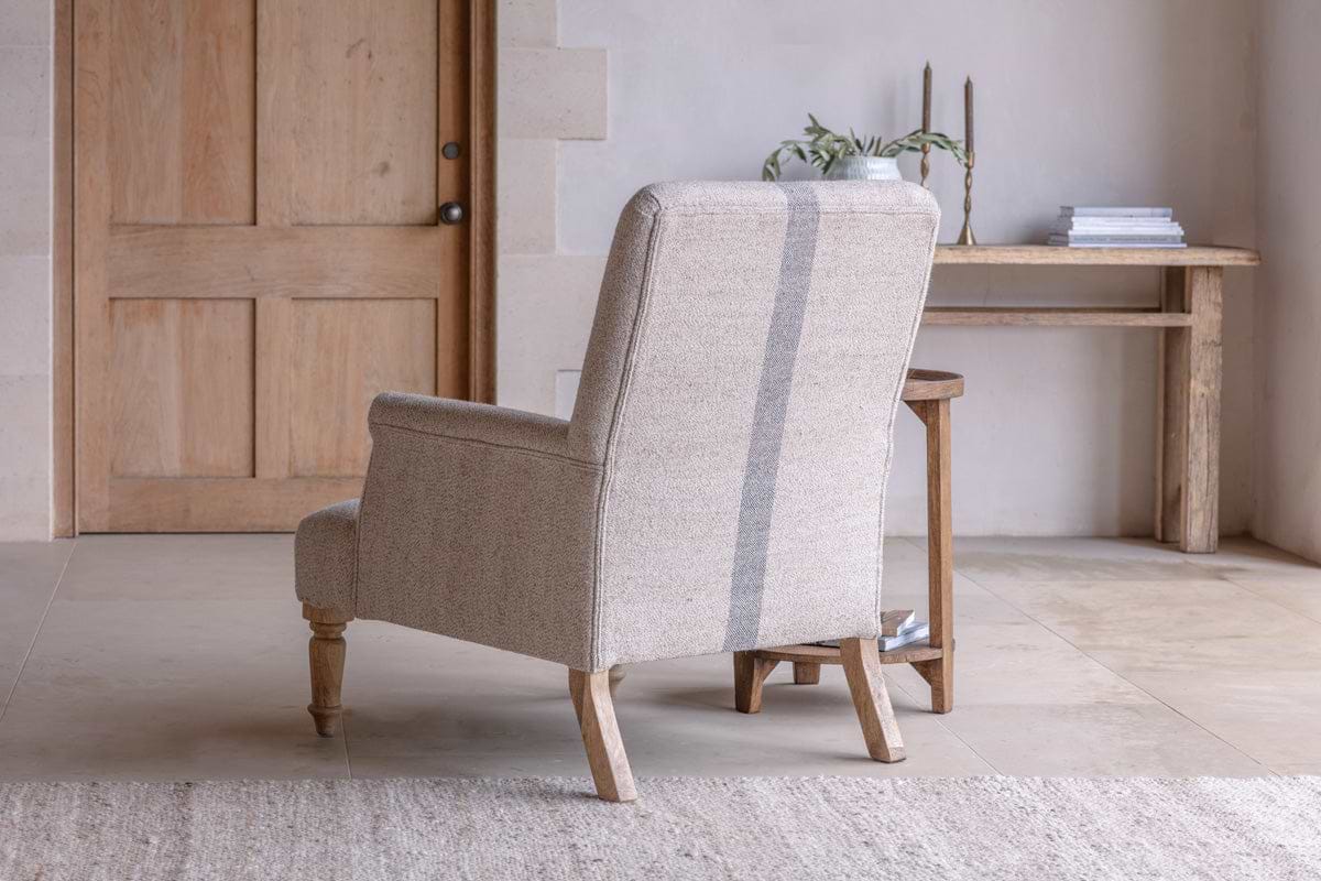 The nkuku Kallu Wool & Cotton Occasional Chair - Natural, with textured beige upholstery and a gray vertical stripe, is set by a rustic wooden door and distressed console table with books and candles, adding charm to any cozy nook.