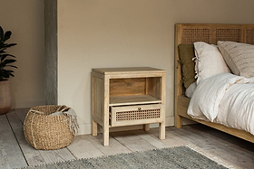A cozy bedroom scene with the nkuku Kanz Mango Wood Bedside Table featuring handwoven cane, an open shelf and drawer, a cane headboard bed with white bedding, woven basket, plant on the left, and gray rug on light wood floor.