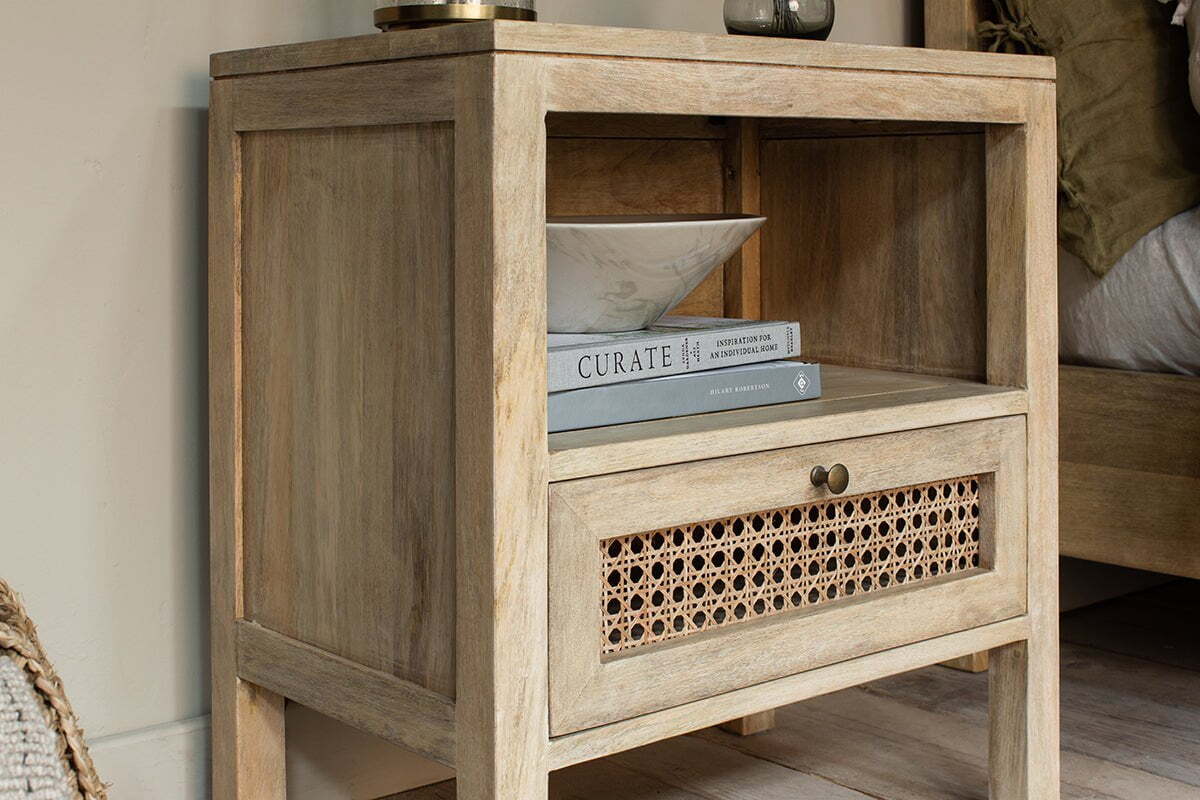 The nkuku Kanz Mango Wood Bedside Table features an open shelf for books and a bowl, plus a drawer with a handwoven cane insert and brass knob, offering stylish and practical bedside storage.