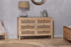 The nkuku Kanz Mango Wood Chest Of Drawers - Natural features six rattan drawers for stylish storage. A glass lamp with burlap shade, brass candlestick, and geometric lantern are on top; a tufted armchair sits left, wooden bed frame right.