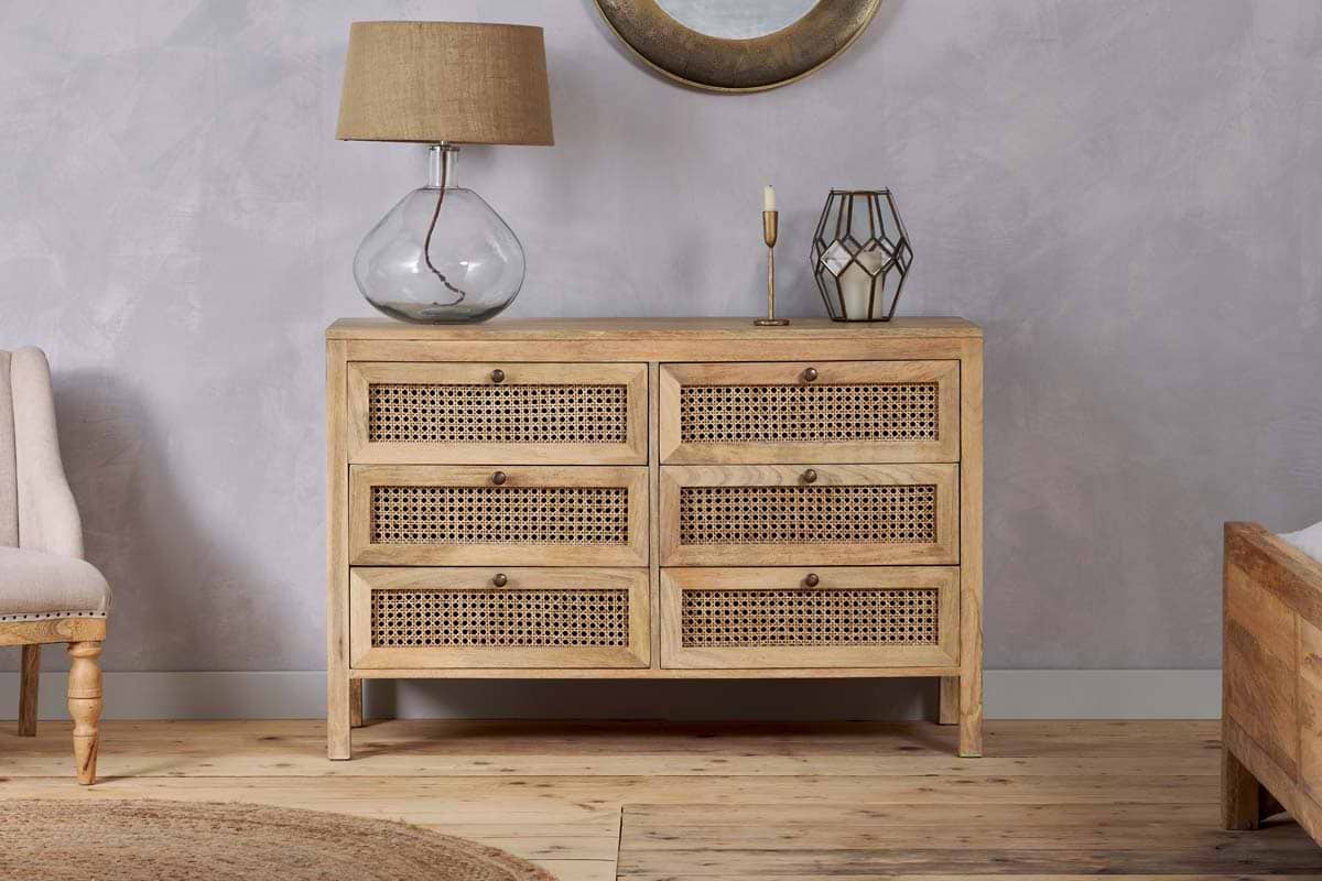 The nkuku Kanz Mango Wood Chest Of Drawers - Natural, featuring six wicker-fronted drawers, stands against a light gray wall. On top are a glass lamp with a tan shade, gold candleholder, and decorative lantern for stylish bedroom storage.