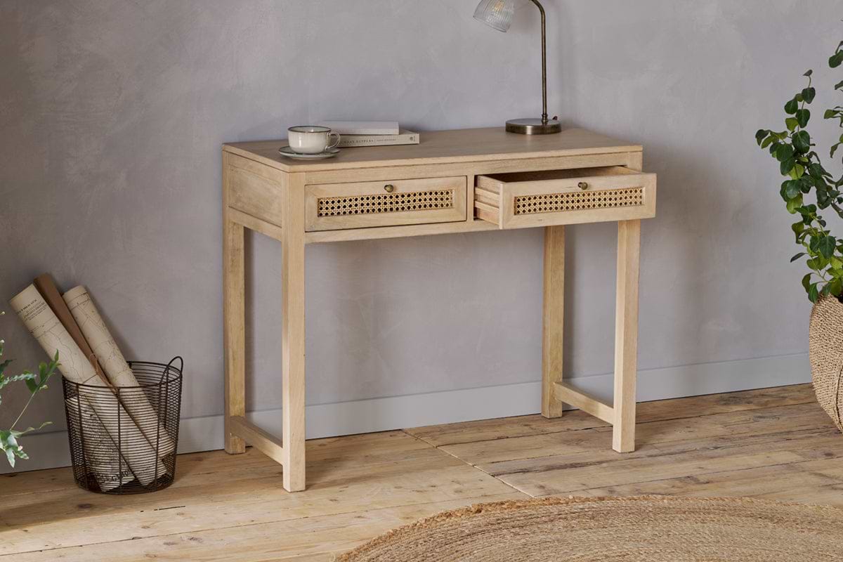 The nkuku Kanz Mango Wood Writing Desk - Natural stands against a gray wall, topped with a lamp, cup, and book. Rolled papers in a wire basket, a braided rug, and green plants accent the floor—ideal home office furniture.