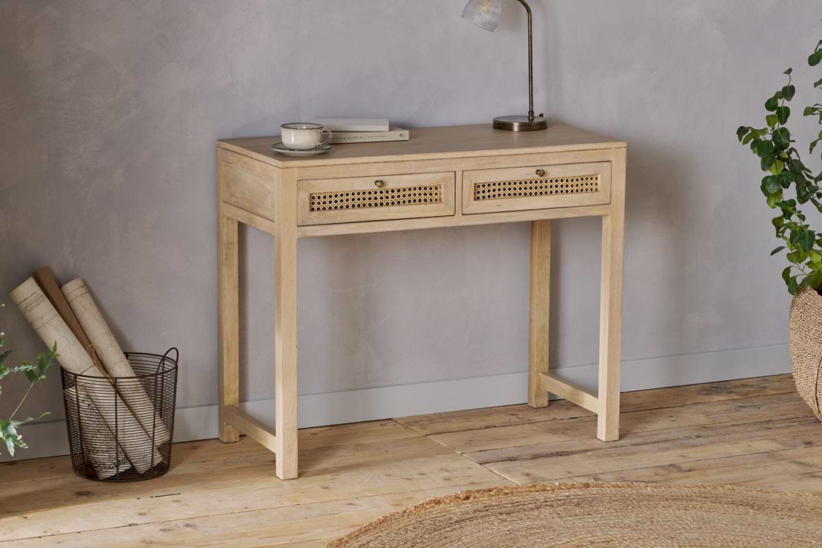 The nkuku Kanz Mango Wood Writing Desk - Natural stands against a gray wall, topped with a book, cup, and lamp. A wire basket with rolled papers sits to the left and green plants to the right. A round woven rug lies on the floor.