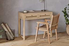 The nkuku Kanz Mango Wood Writing Desk - Natural stands against a gray wall, paired with a matching chair. Rolled papers, a plant, and a round rug on the wooden floor complete this cozy home office setup.