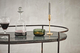 A glass of red wine, a decanter, olives in a green bowl, and a tall silver candle in a gold holder adorn the nkuku Karoko Marble Drinks Table, bringing elegance to any gathering against a textured light gray wall.