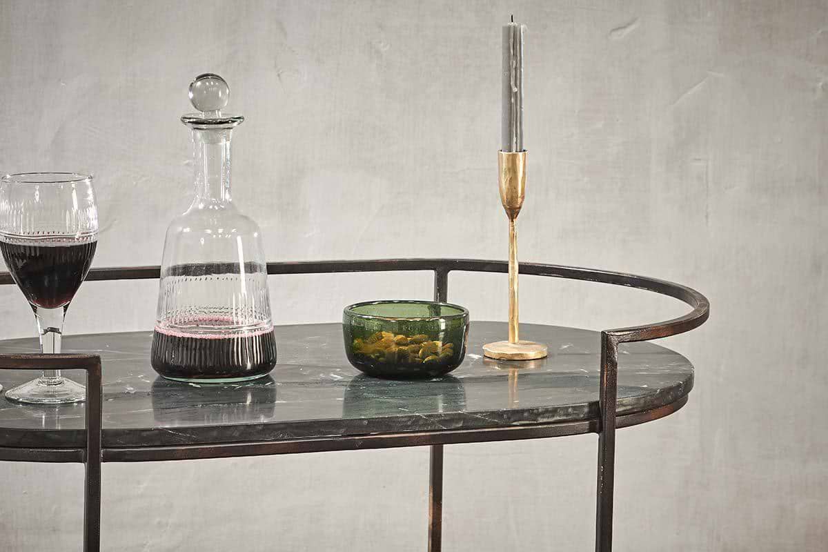 A glass of red wine, a decanter, olives in a green bowl, and a tall silver candle in a gold holder adorn the nkuku Karoko Marble Drinks Table, bringing elegance to any gathering against a textured light gray wall.