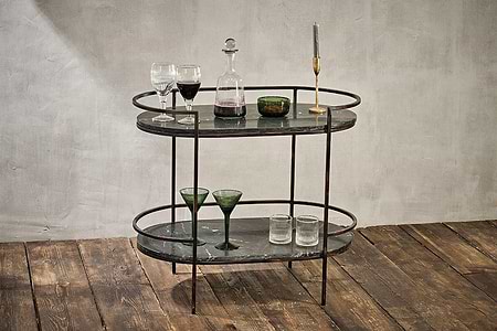 The Karoko Marble Drinks Table by nkuku features two oval black marble shelves and a distressed metal frame, perfect for displaying glassware, a decanter of red wine, a green bowl, and a gold candlestick with a grey candle.
