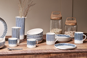 A rustic wooden table showcases nkuku’s Karuma Ceramic Mug - Large (Set of 2), featuring white ceramic with blue stripes, alongside matching bowls, plates, and a vase with dried branches. Two large glass jars with wooden lids in the background hold grains or cereal.