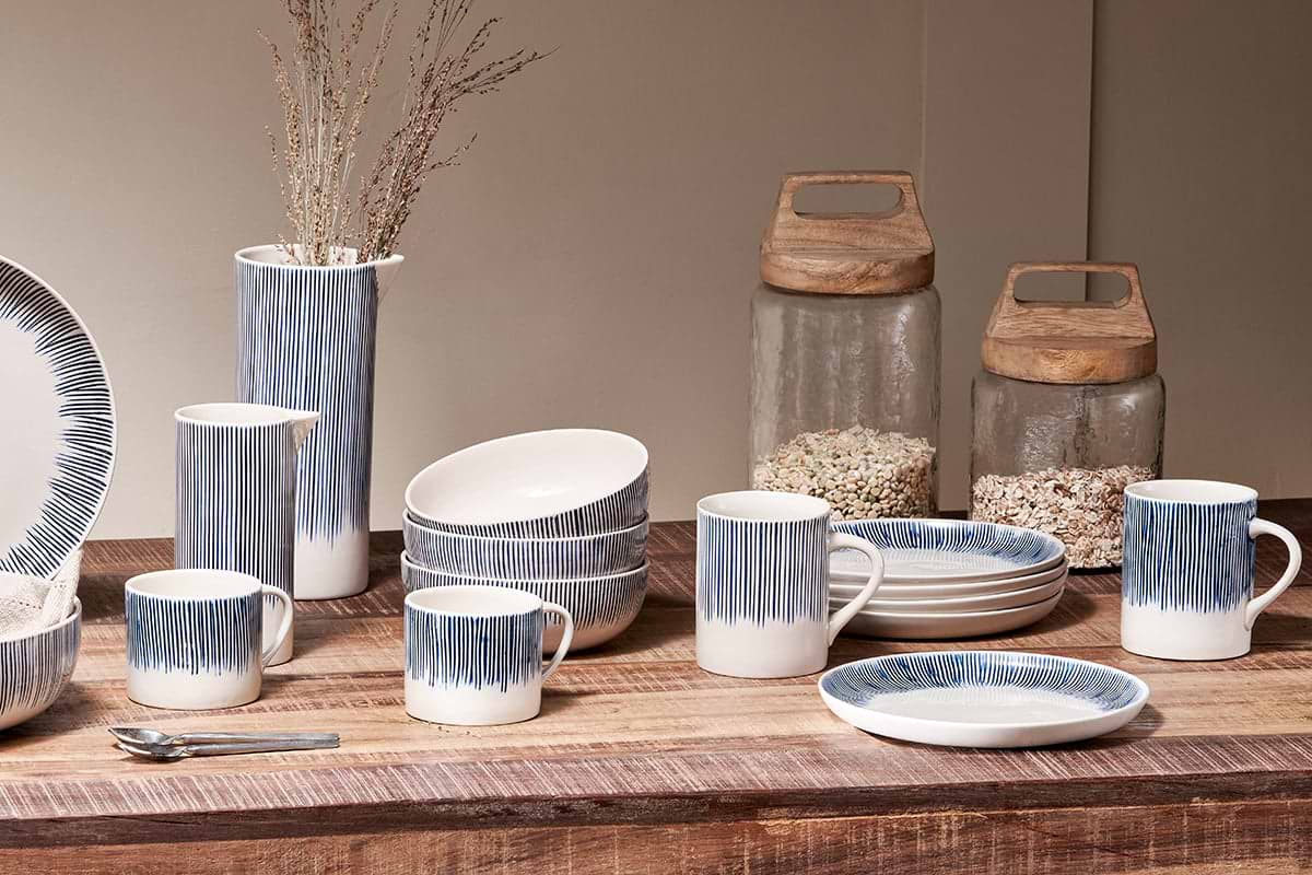 A rustic wooden table showcases nkuku’s Karuma Ceramic Mug - Large (Set of 2), featuring white ceramic with blue stripes, alongside matching bowls, plates, and a vase with dried branches. Two large glass jars with wooden lids in the background hold grains or cereal.
