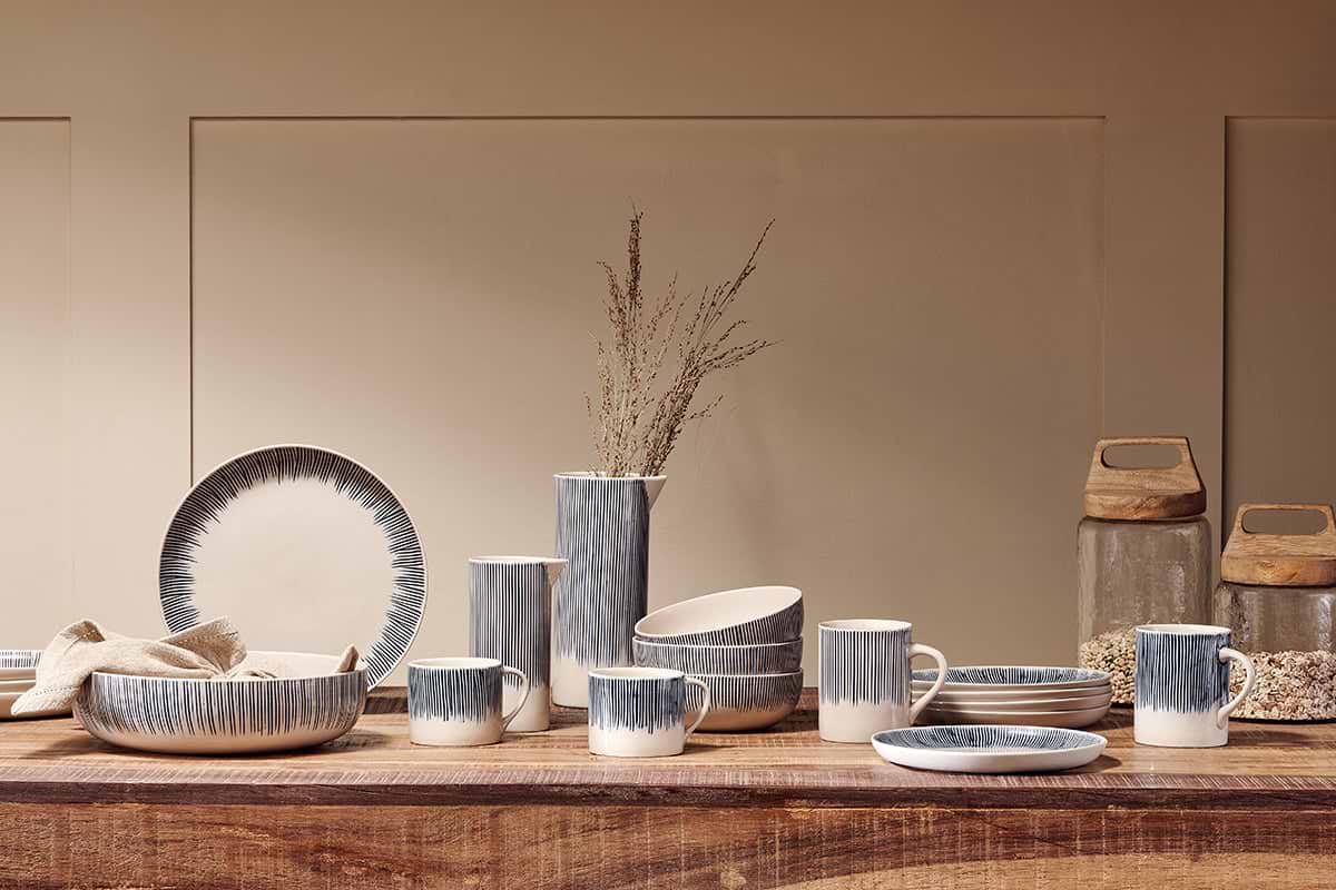 A wooden table showcases nkuku’s Karuma Ceramic Mug - Large (Set of 2) alongside handmade bowls, dishes, and a vase with dried grass. The blue-gray and white gradients create a modern rustic style, with two jars of grains nearby against a beige wall.
