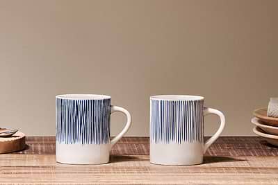 Two white nkuku Karuma Ceramic Mug - Large (Set of 2), featuring vertical blue lines, sit side by side on a wooden surface, with handmade mugs, stacked plates, a napkin, and a cork coaster in the neutral beige background.
