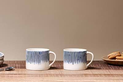 A set of two Karuma Ceramic Mugs - Small by nkuku, featuring blue vertical stripes, are placed on a wooden table with a spoon to the left and a small bowl of cookies to the right.