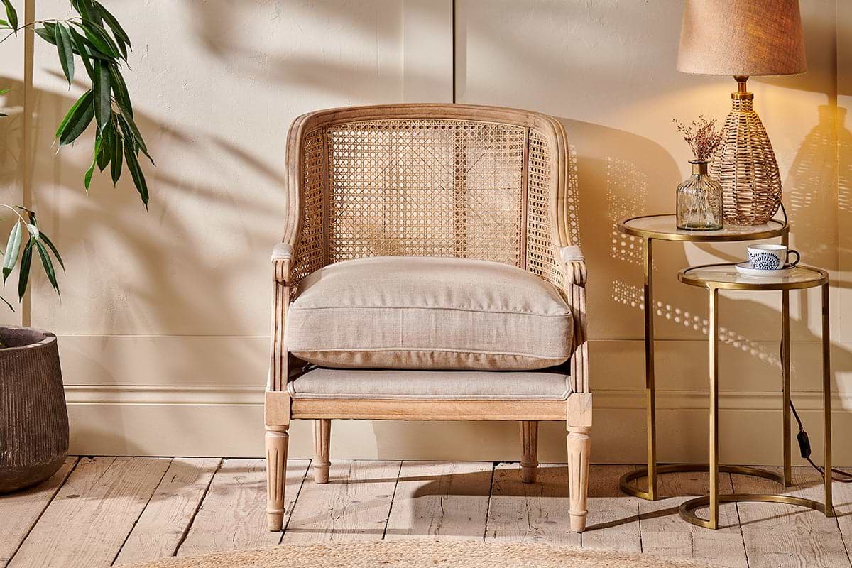 A cozy room features the Kaziria Cane & Linen Armchair - Natural by nkuku with a beige cushion. Nearby, a round side table holds a lamp, glass bottle, and coffee cup. A potted plant sits left as sunlight casts shadows on the cream wall and wood floor.