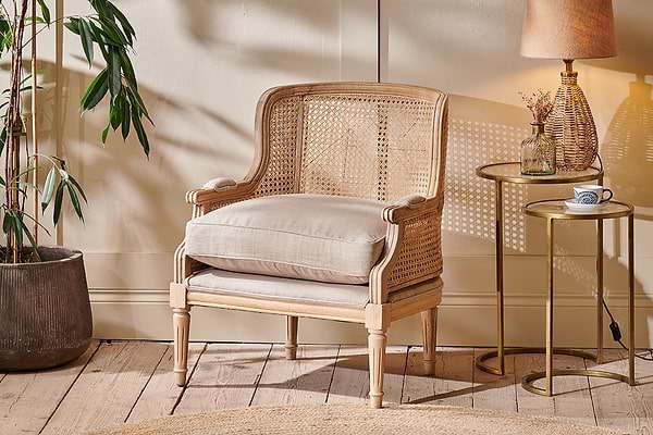 The nkuku Kaziria Cane & Linen Armchair - Natural, featuring a light wood frame and neutral upholstery, sits beside a side table topped with a lamp, cup, and vase.