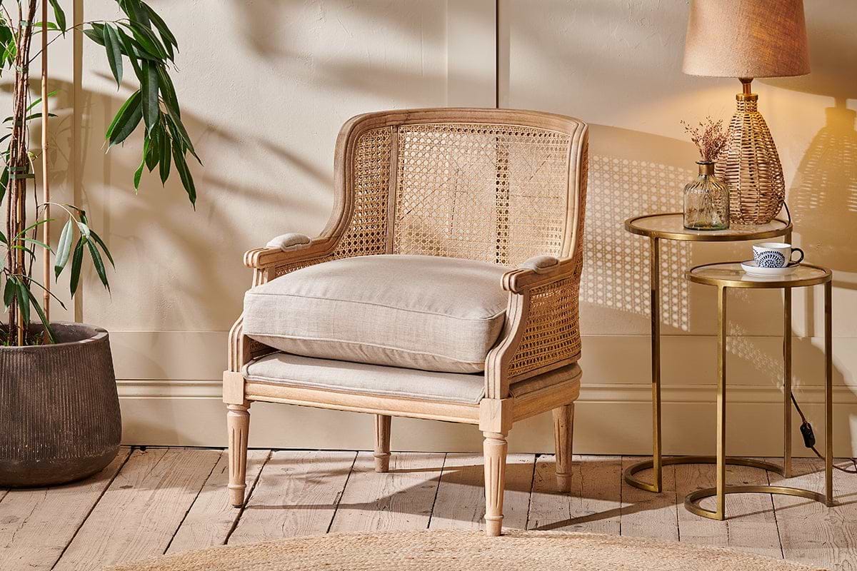 The nkuku Kaziria Cane & Linen Armchair - Natural, featuring a light wood frame and neutral upholstery, sits beside a side table topped with a lamp, cup, and vase.
