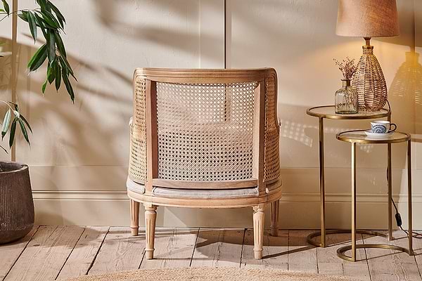 The Kaziria Cane & Linen Armchair by nkuku, with its natural wooden frame, sits on a rustic floor beside a round side table holding a lamp, vase, and cup. A leafy plant and soft natural light enhance the room's cozy, neutral tones.