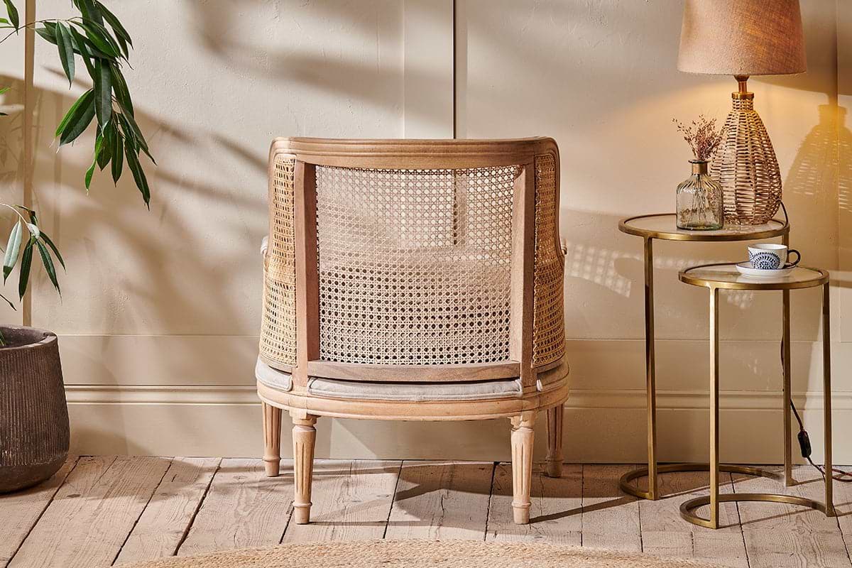 The Kaziria Cane & Linen Armchair by nkuku, with its natural wooden frame, sits on a rustic floor beside a round side table holding a lamp, vase, and cup. A leafy plant and soft natural light enhance the room's cozy, neutral tones.