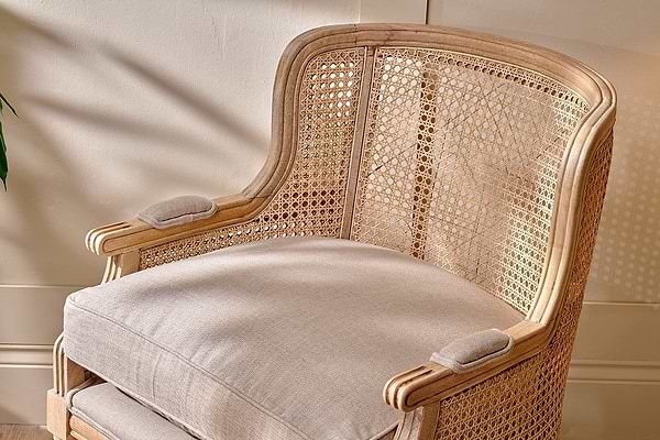 The Kaziria Cane & Linen Armchair by nkuku features natural linen, a light mango wood frame, and woven cane back and sides. Its beige cushion and padded armrests offer comfort, perfect for any space with a wooden floor and cream wall.